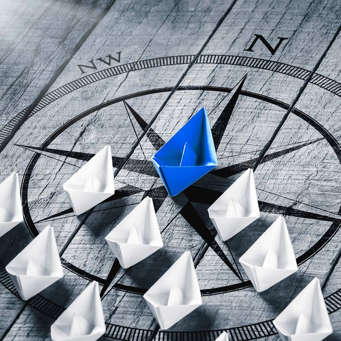 Paper boats on a compass, one blue leading white boats, symbolizing leadership and direction.