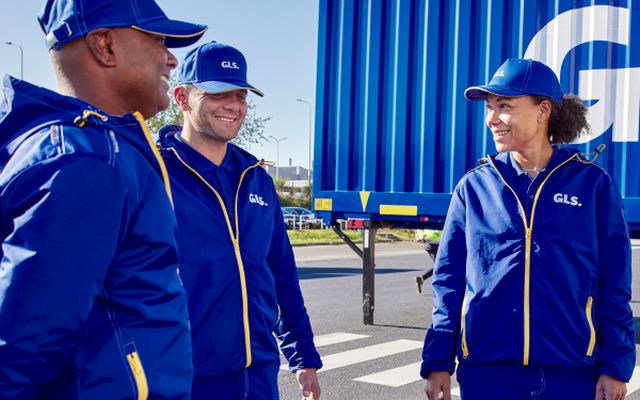Three GLS employees in blue uniforms smiling and talking outdoors near a blue shipping container.