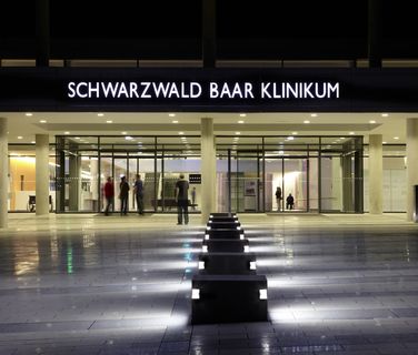 Modern hospital entrance at night with SCHWARZWALD BAAR KLINIKUM sign, people visible inside.