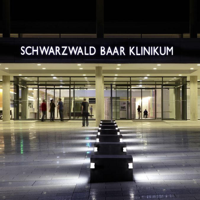 Modern hospital entrance at night with SCHWARZWALD BAAR KLINIKUM sign, people visible inside.