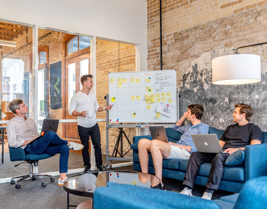 Team meeting in modern office, discussing tasks on whiteboard; laptops open, collaborative workspace.