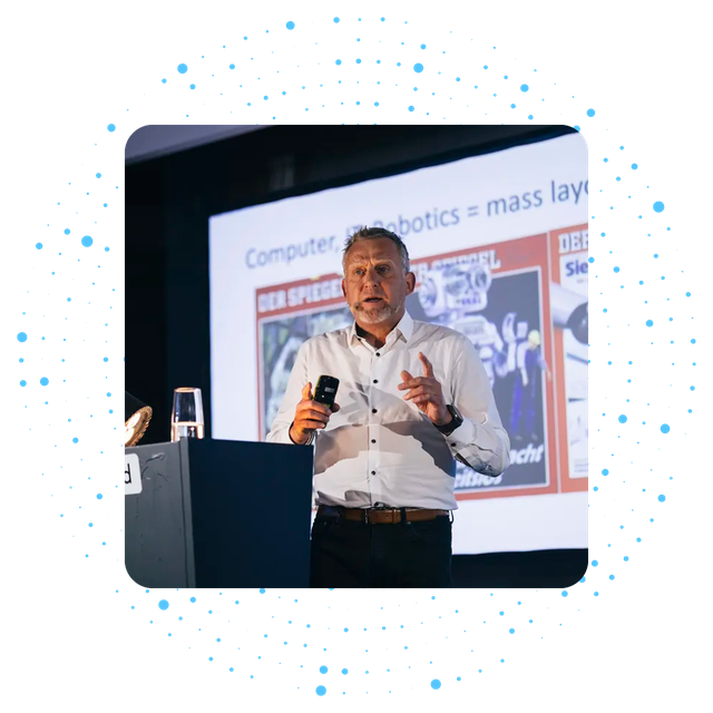 Man presenting on stage with a slide about robotics and mass layoffs.