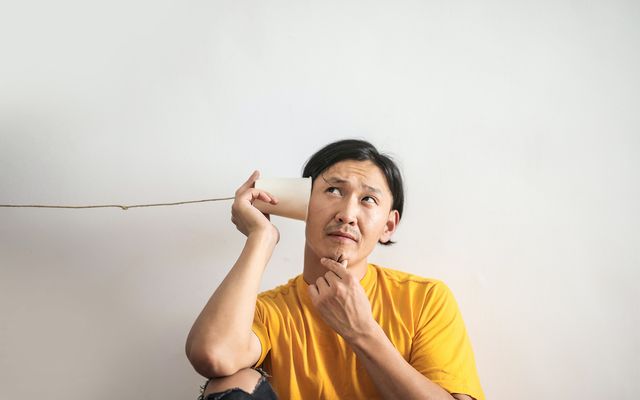 Man in yellow shirt listening to a tin can phone, thinking, on plain background.