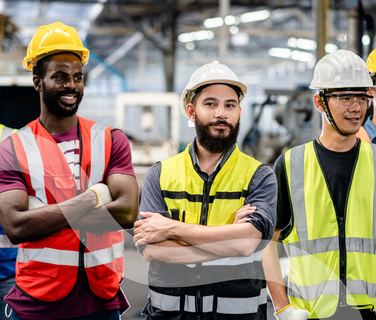 Image of three manufacturing workers in hard hats and high-vis vests, with one staring down the lens with crossed arms. All look happy.