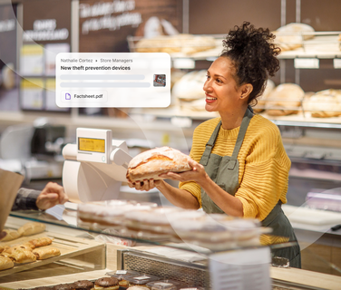 A smiling bakery employee in a yellow sweater and apron holds a loaf of bread behind the counter, with various baked goods displayed.