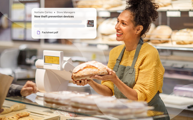 A smiling bakery employee in a yellow sweater and apron holds a loaf of bread behind the counter, with various baked goods displayed.