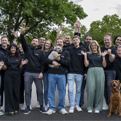 A group of people smiling and waving, with a dog and a puppy, outdoors in a park setting. They are wearing matching black hoodies.