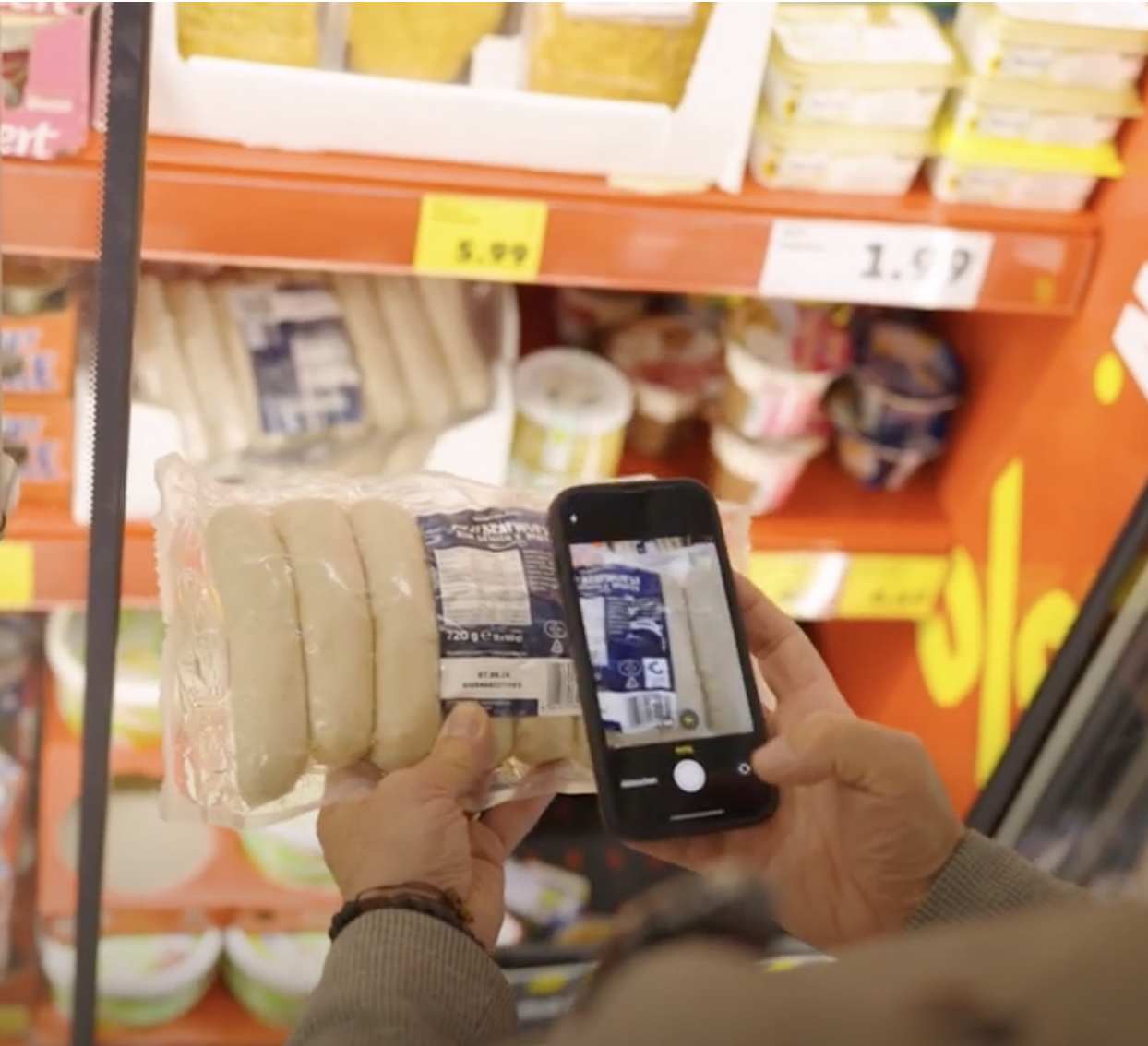 Person scans packaged sausages with smartphone in supermarket; price tags show 5.99 and 1.59.