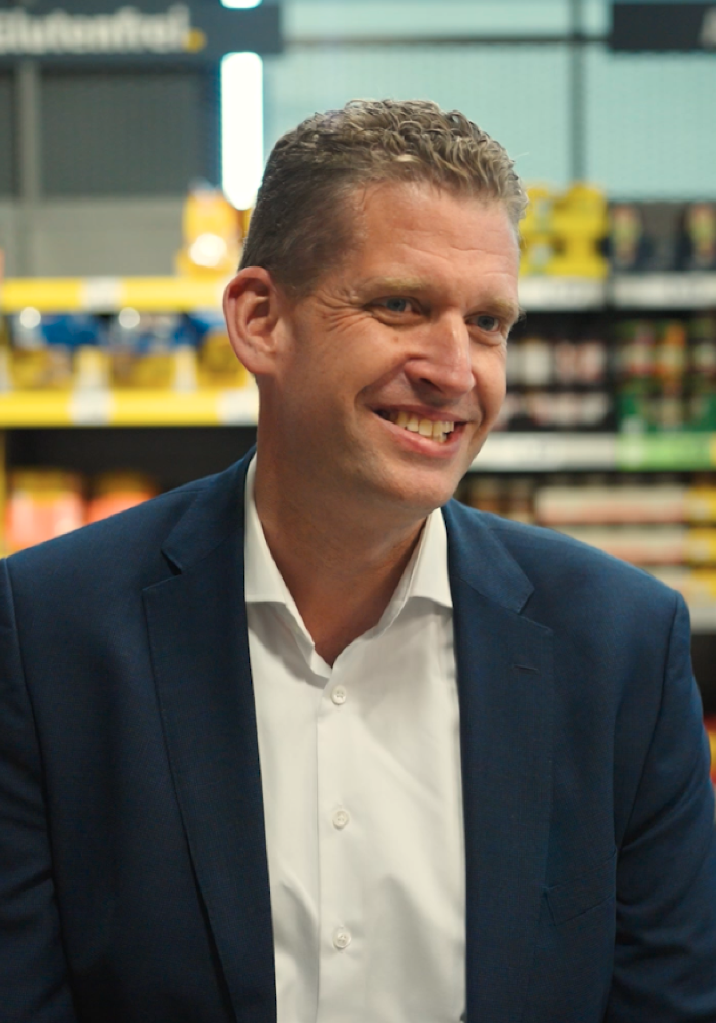Smiling man in business attire in a supermarket aisle, glutenfrei sign visible in background.