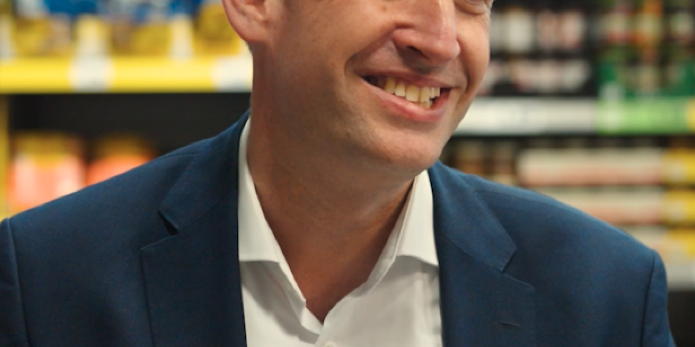 Smiling man in business attire in a supermarket aisle, glutenfrei sign visible in background.