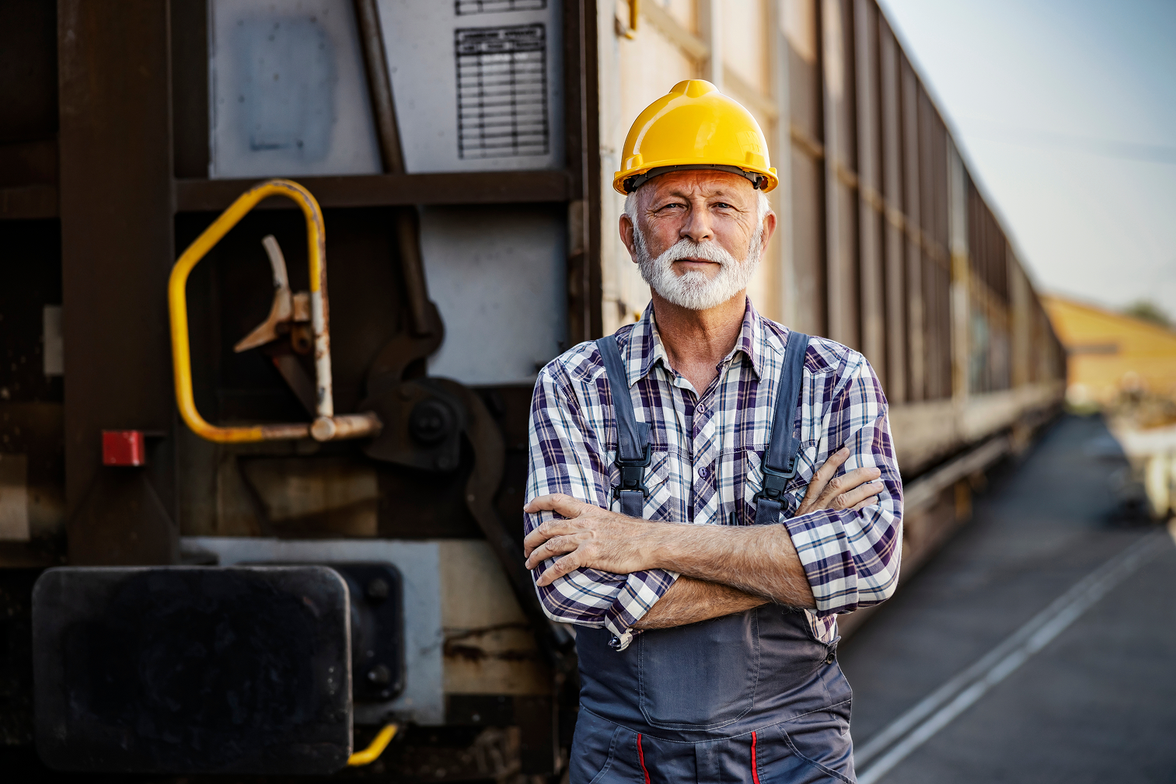 Older male worker in hard hat and overalls stands confidently by a train, industrial background.
