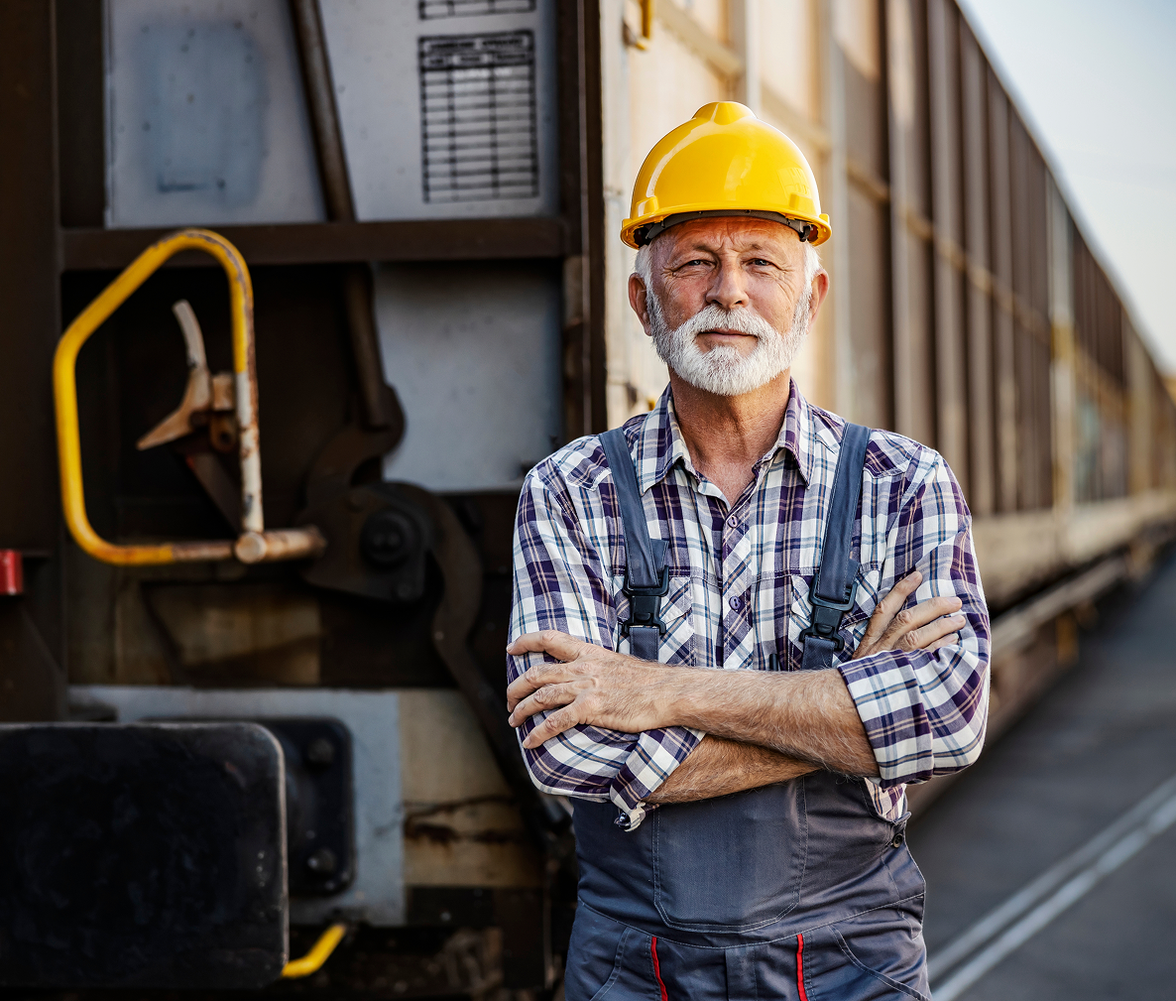 Older male worker in hard hat and overalls stands confidently by a train, industrial background.