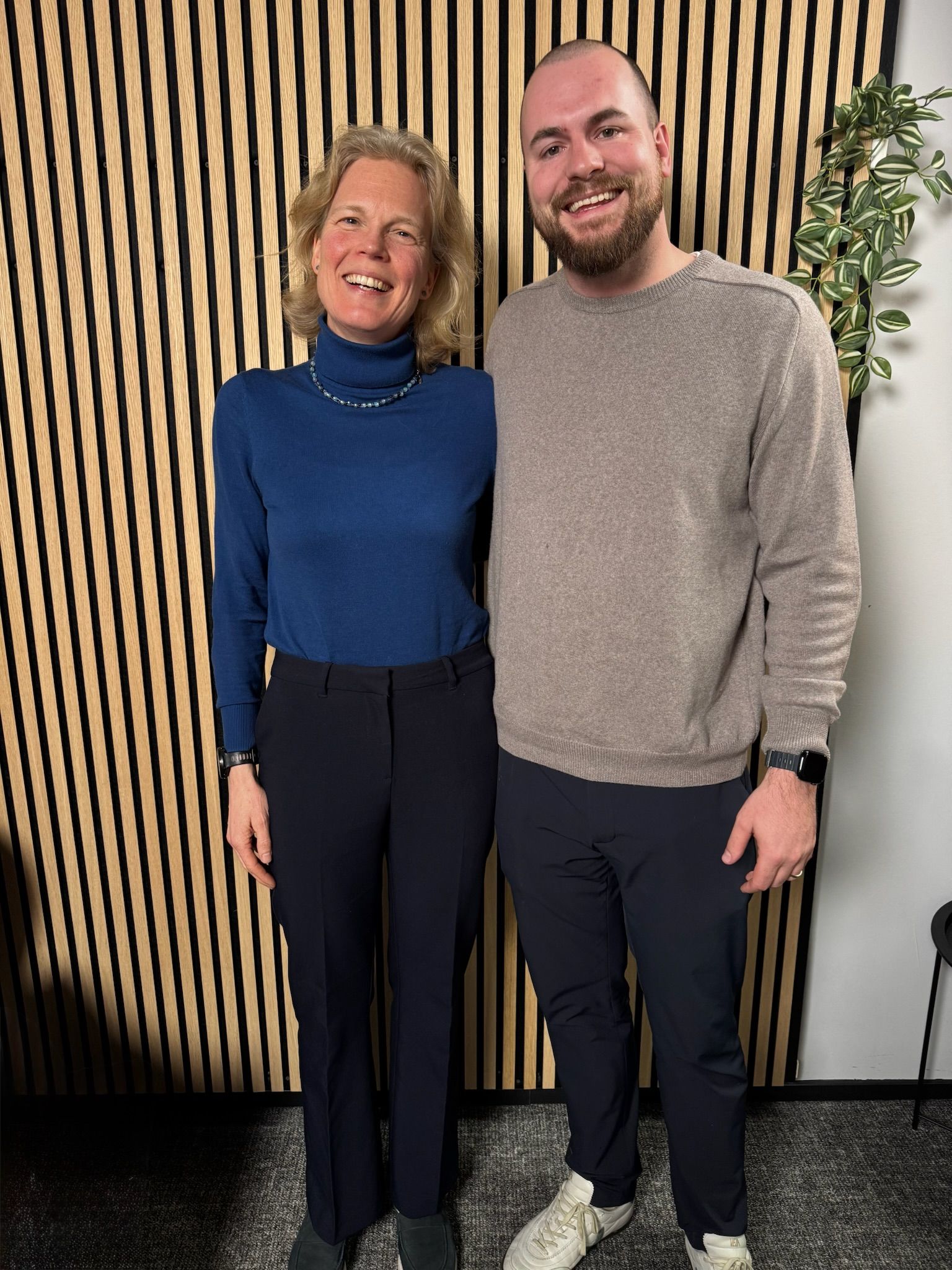 Two people smiling in front of a wooden panel wall.