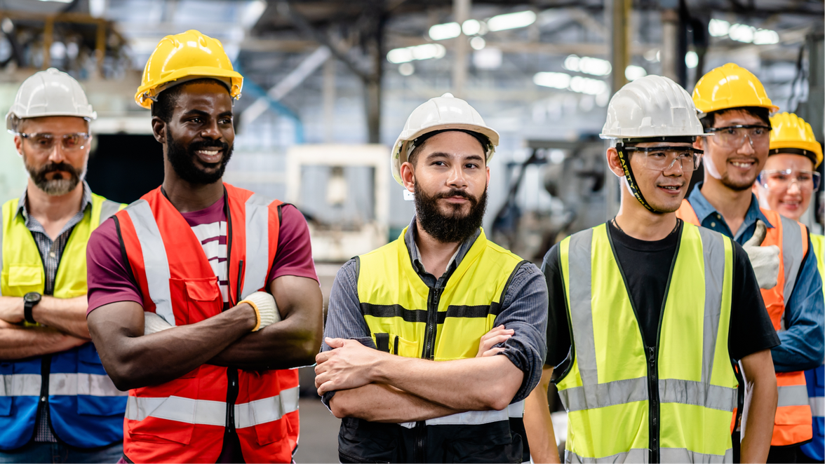 A diverse group of construction workers wearing safety vests and helmets stand confidently in an industrial setting.