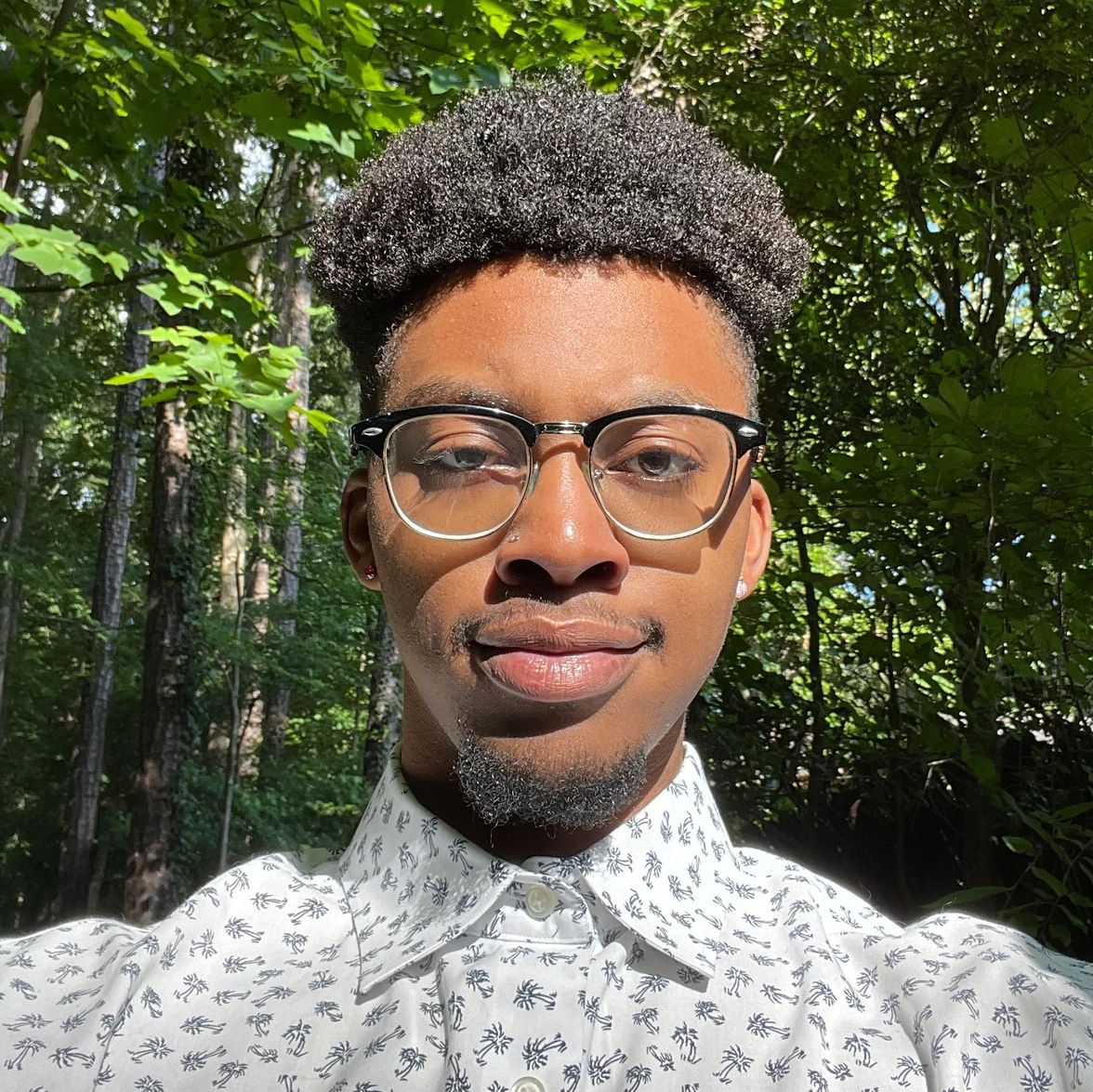 A person wearing glasses and a patterned shirt stands outdoors in a sunlit, leafy forest.