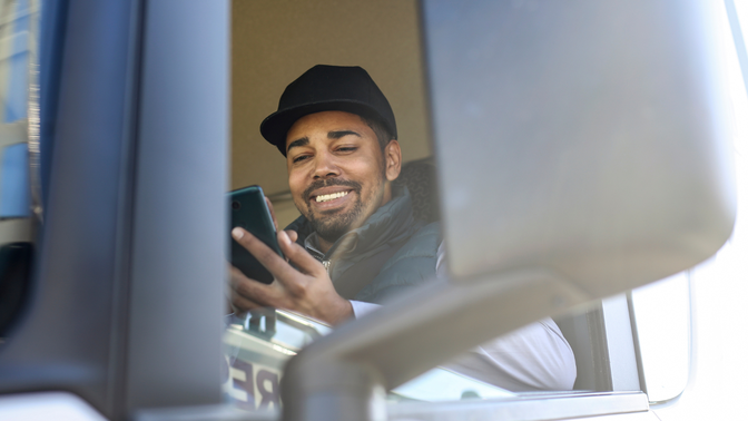 Smiling man in a cap looking at his phone while sitting in the driver's seat of a vehicle.