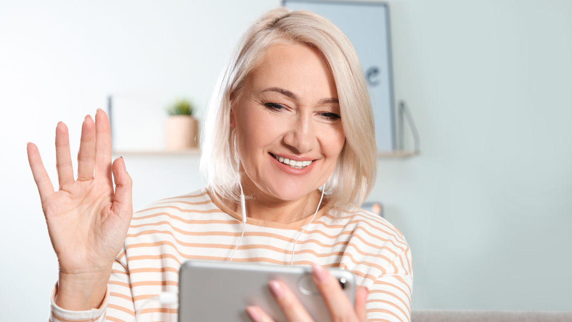 A smiling woman with short blonde hair waves at her smartphone during a video call, wearing earbuds and seated in a bright, home-like setting.
