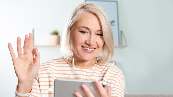 A smiling woman with short blonde hair waves at her smartphone during a video call, wearing earbuds and seated in a bright, home-like setting.