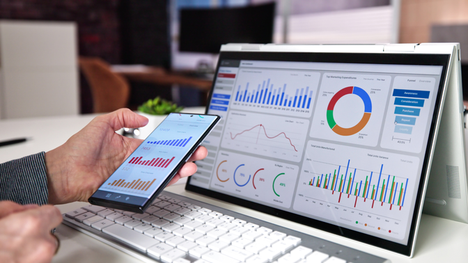 Person analyzing graphs on a tablet and laptop with colorful charts, bar graphs, and pie charts in a modern office setting.