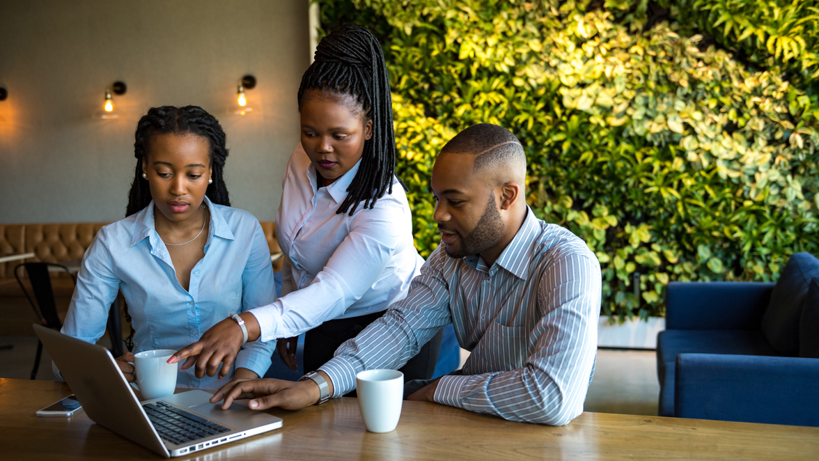 Three employees collaborate at a laptop in a modern office, discussing workforce management scheduling.
