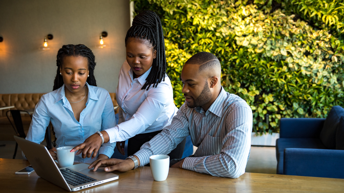 Three people in a modern office setting collaborate at a laptop. Two women and a man, all with coffee mugs, focus on the screen.