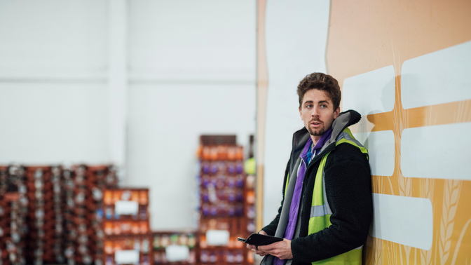 A warehouse worker in a high-visibility vest leans against a wall, holding a tablet and looking off-camera, with pallets in the background.