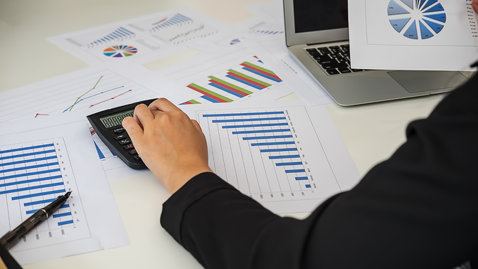 Person analyzing printed graphs and charts at a desk, using a calculator and laptop.