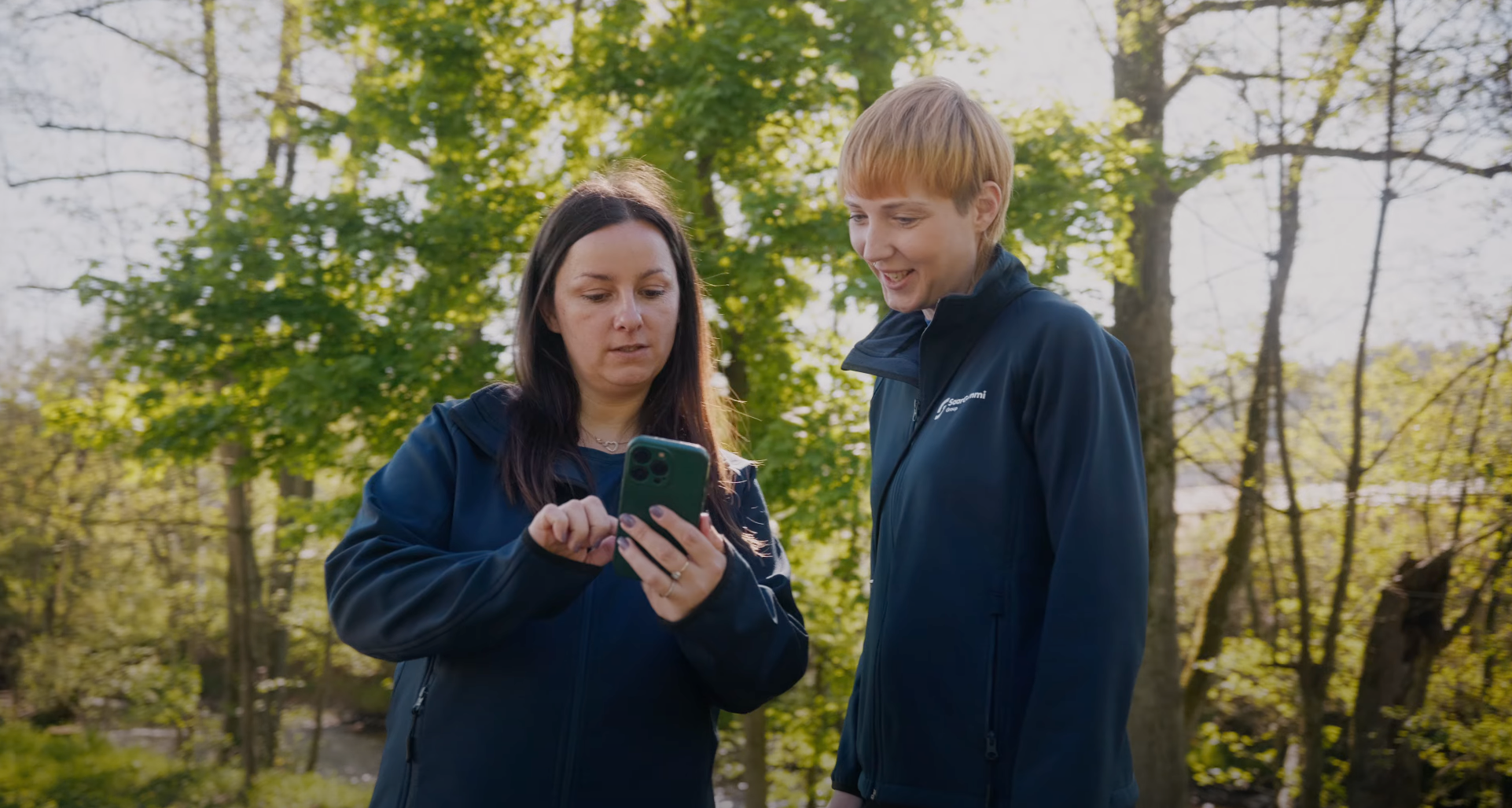 Two people outdoors in branded jackets, one showing something on a smartphone to the other.
