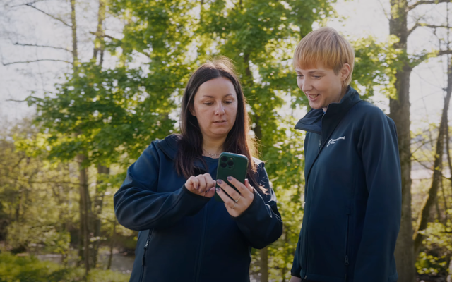 Two people outdoors in branded jackets, one showing something on a smartphone to the other.