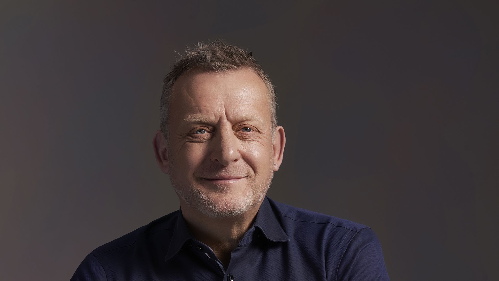 Portrait of a man in a dark shirt sitting on a stool, neutral background, no visible text.