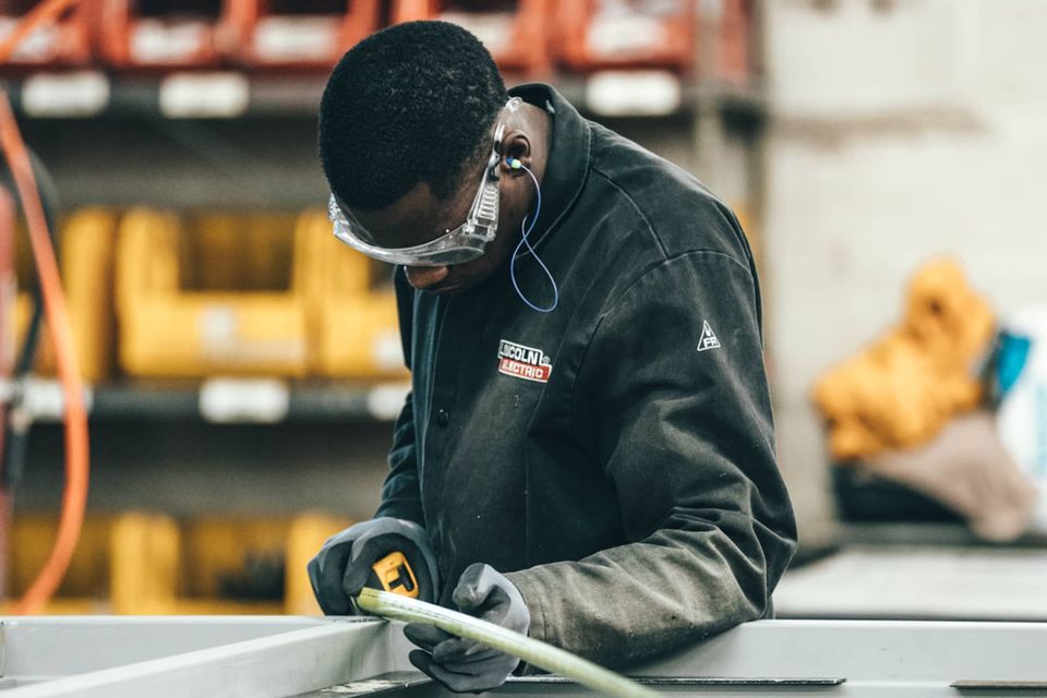 Manufacturing worker measures metal with tape measure, wearing safety goggles and gloves in workshop.
