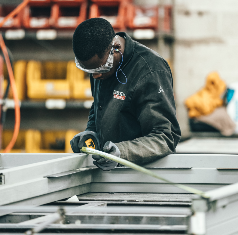 A worker in safety gear measures metal with a tape measure in an industrial workshop setting.