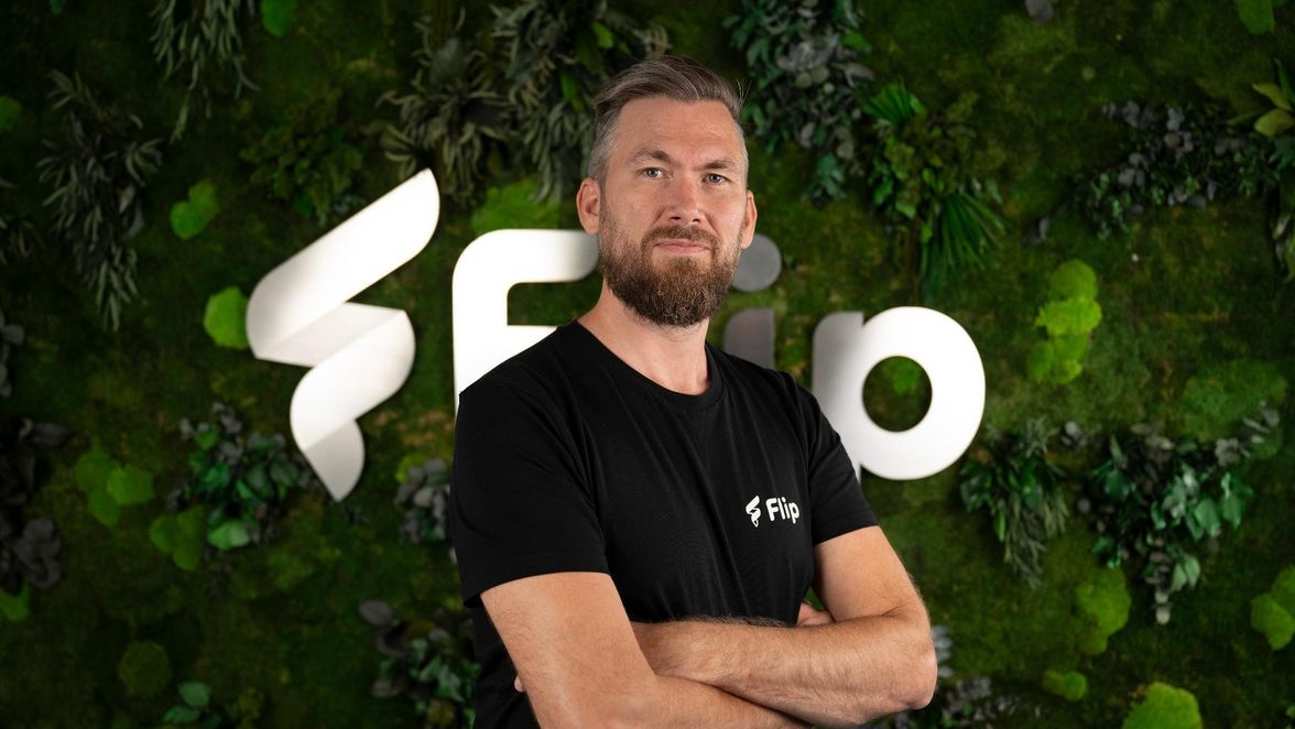 Man in black Flip shirt poses in front of green wall with large Flip logo, no UI visible.