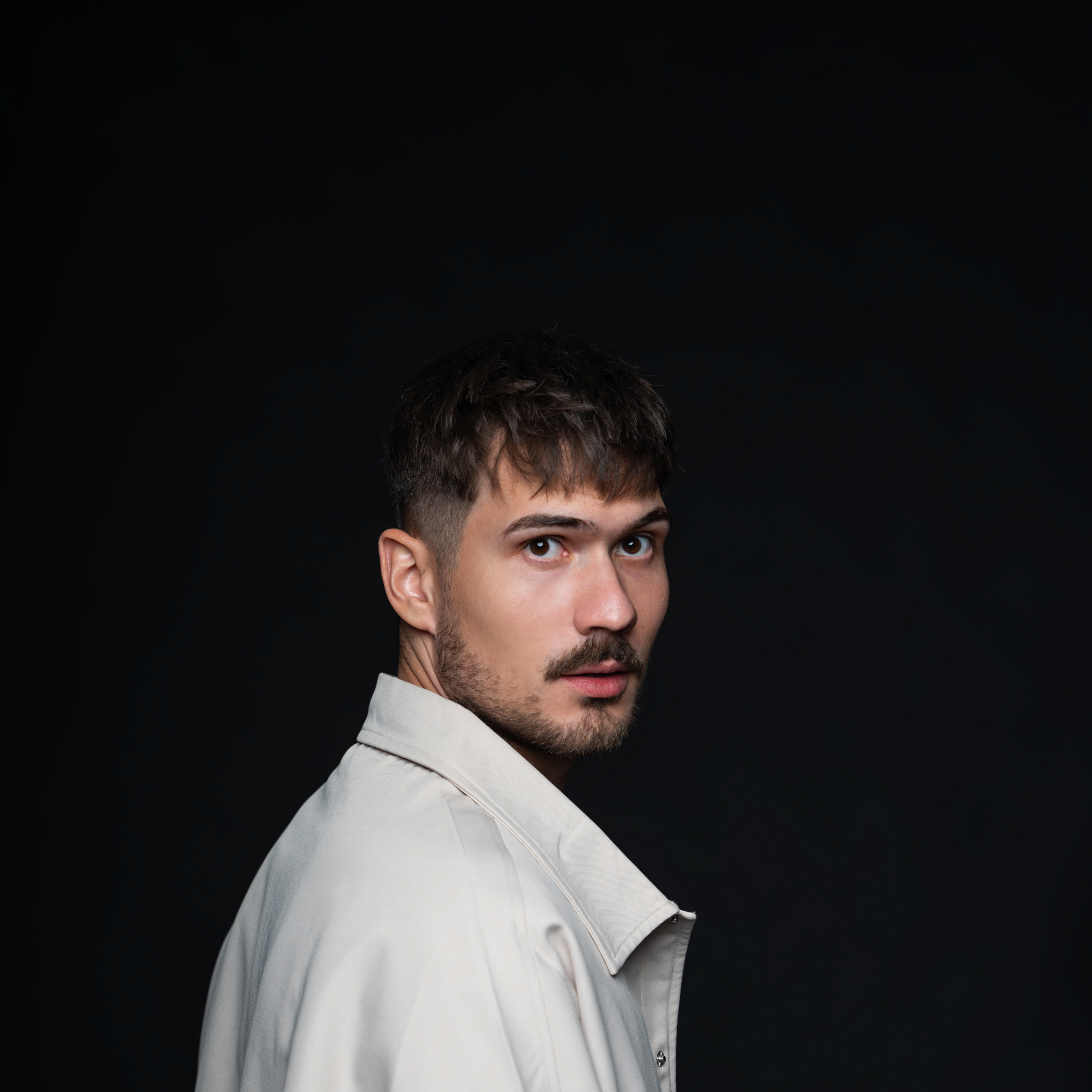 A man in a light jacket poses against a dark background, looking over his shoulder.