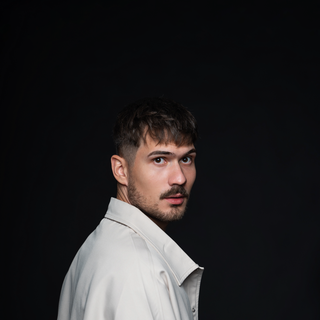 A man in a light jacket poses against a dark background, looking over his shoulder.