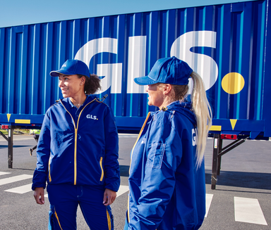Two GLS employees in blue uniforms stand and talk in front of a GLS-branded shipping container.