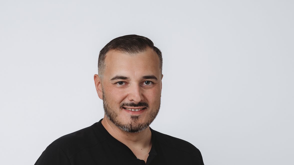 Man in black t-shirt and jeans standing against plain white background, one hand in pocket.
