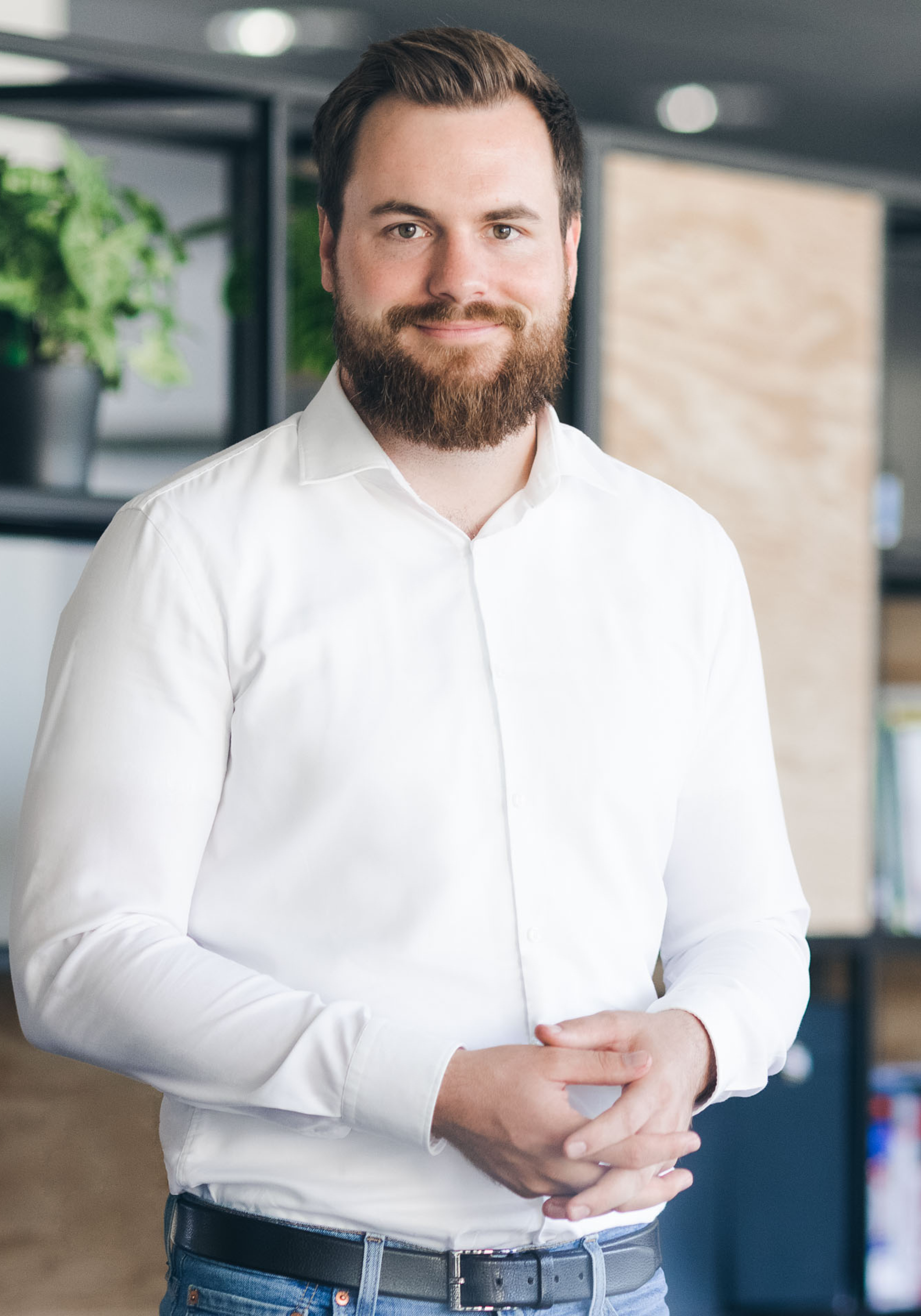 Smiling man in white shirt stands in modern office with plants and bookshelves in background.