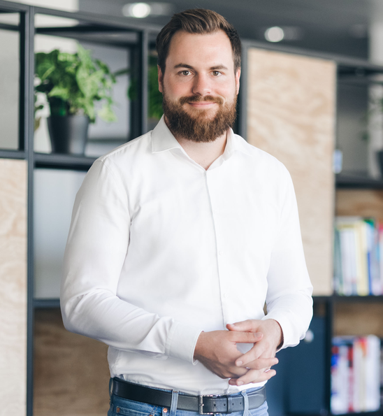 Smiling man in white shirt stands in modern office with plants and bookshelves in background.