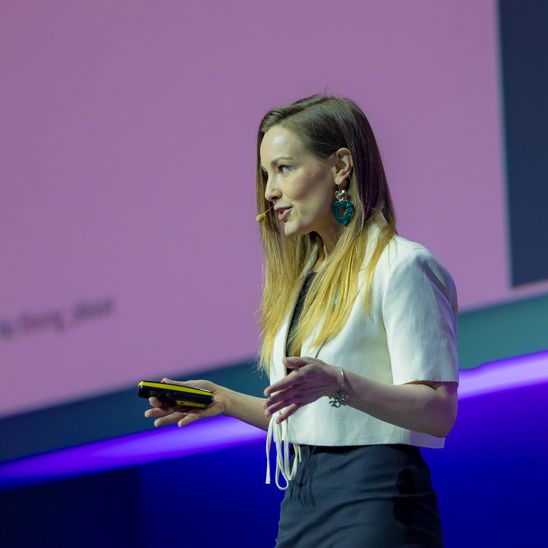 Woman speaking on stage with a headset, holding a device, blurred text in background.