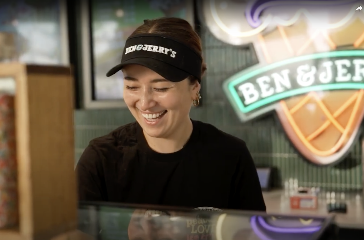 Smiling Ben & Jerrys employee behind counter, wearing branded visor, neon Ben & Jerrys sign behind.