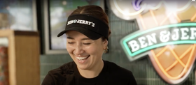 Smiling Ben & Jerrys employee behind counter, wearing branded visor, neon Ben & Jerrys sign behind.