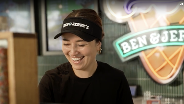 Smiling Ben & Jerrys employee behind counter, wearing branded visor, neon Ben & Jerrys sign behind.