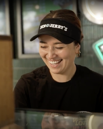 Smiling Ben & Jerrys employee behind counter, wearing branded visor, neon Ben & Jerrys sign behind.