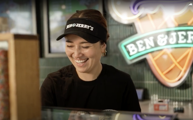 Smiling Ben & Jerrys employee behind counter, wearing branded visor, neon Ben & Jerrys sign behind.