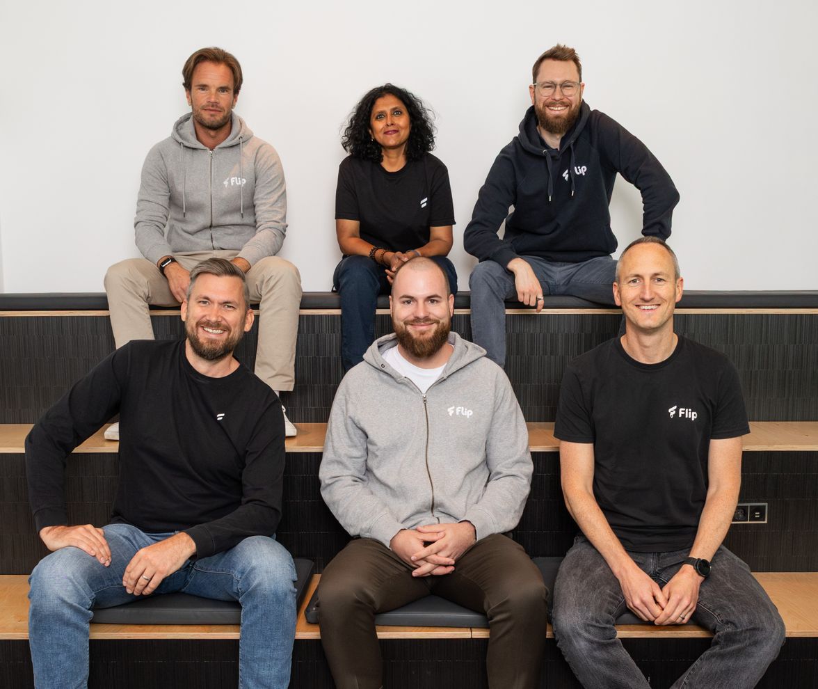 Six smiling Flip management team members in branded shirts, seated on steps, casual office setting.