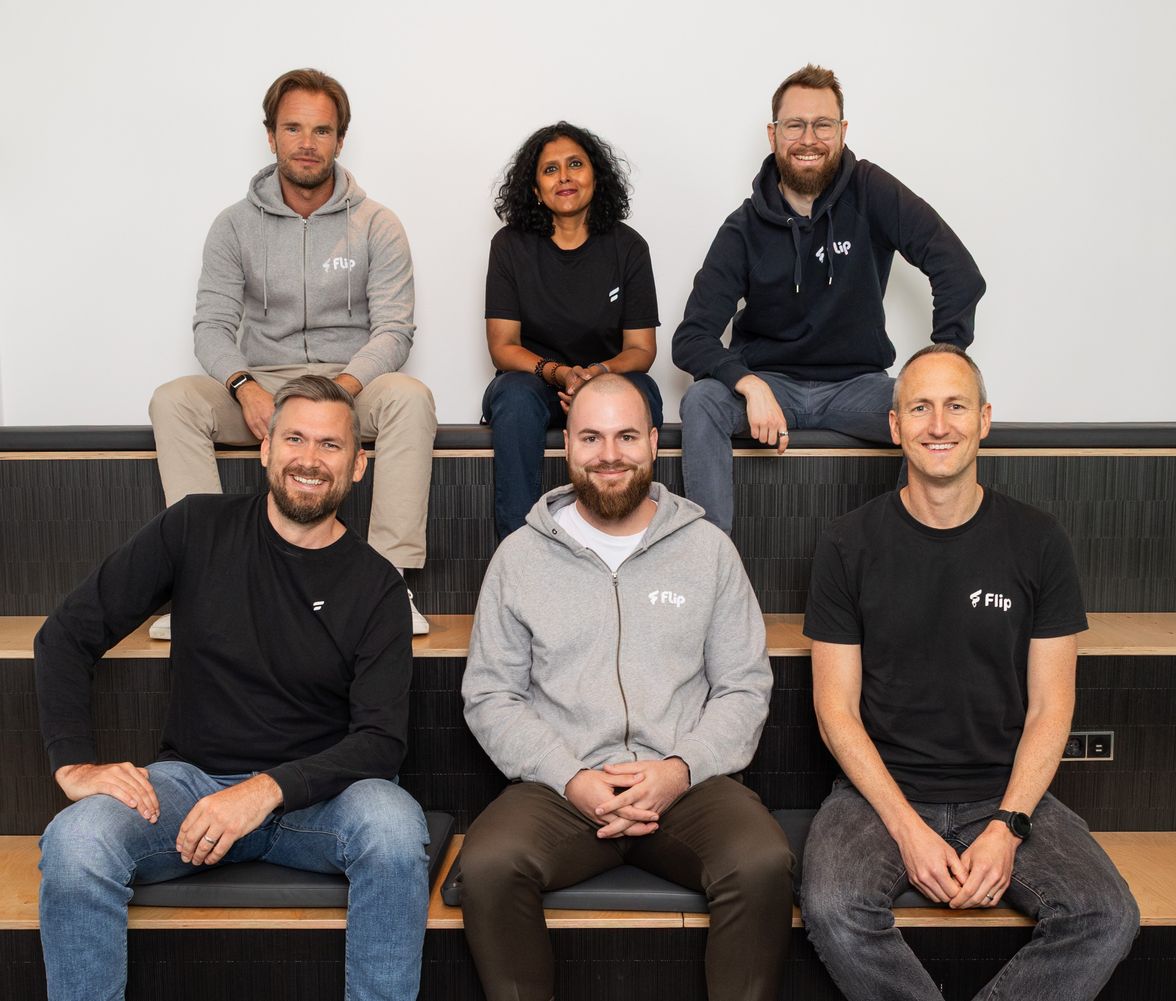 Six smiling Flip management team members in branded shirts, seated on steps, casual office setting.