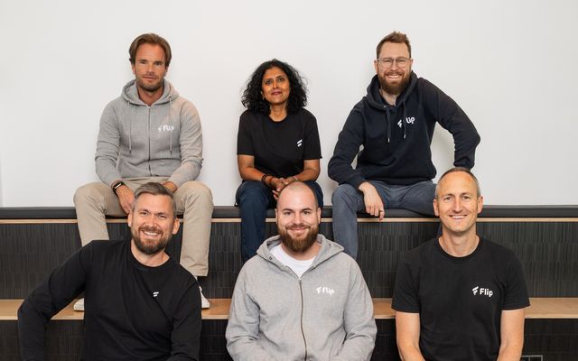 Six smiling Flip management team members in branded shirts, seated on steps, casual office setting.