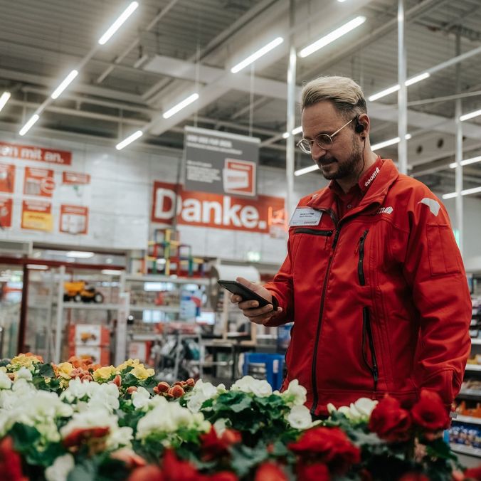 Mitarbeiter in roter Jacke mit Smartphone in einem Baumarkt, umgeben von Blumen.