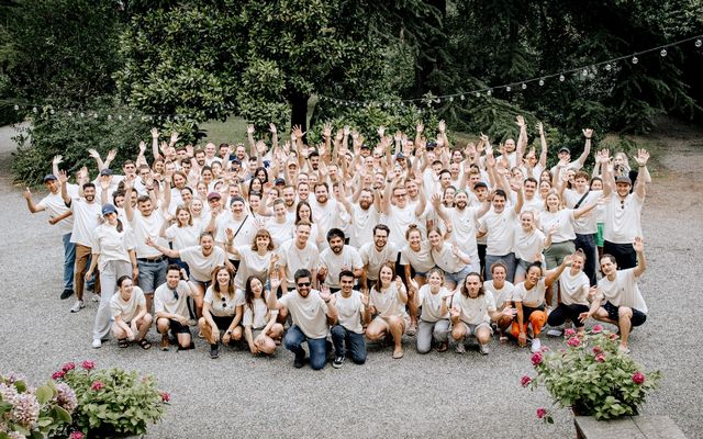 Large group of people in matching shirts outdoors, smiling and waving, team culture event atmosphere.